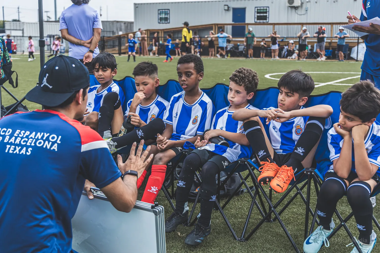 Coach in cap kneeling and talking tactics to seated U10 squad on the sideline