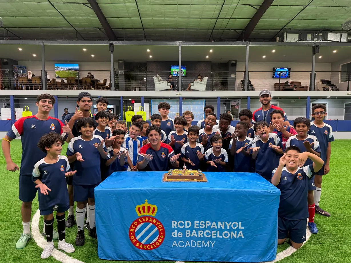 The full RCDE Academy Texas squad celebrating the academy anniversary around a cake on the indoor turf, with the RCD Espanyol de Barcelona Academy banner