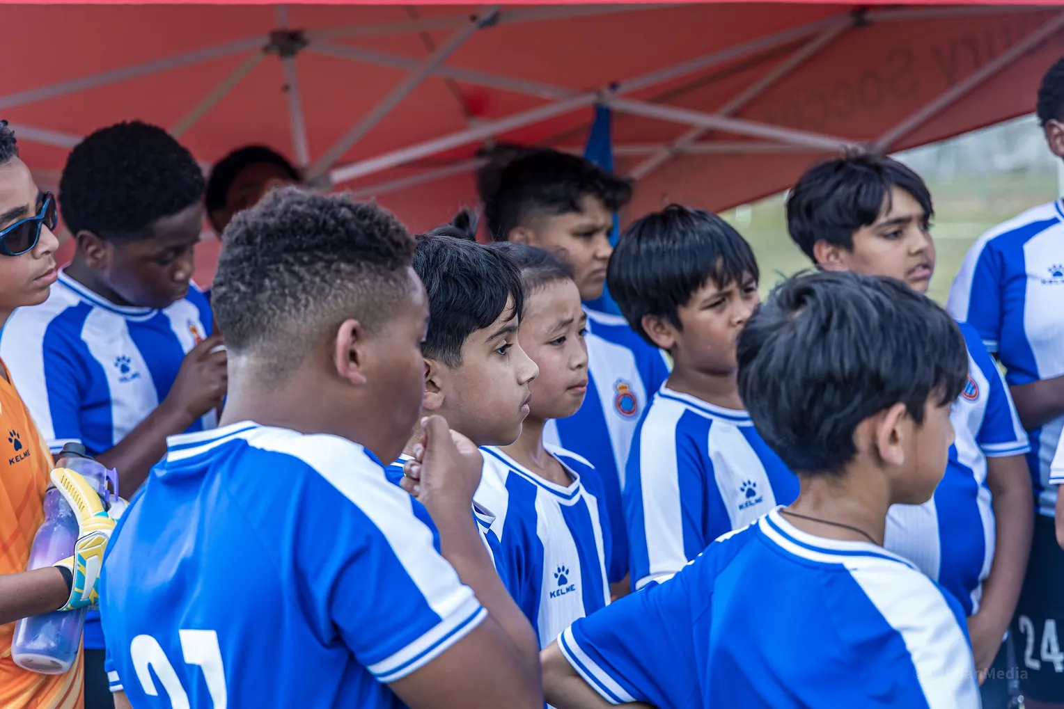 Squad of young academy players gathered under a red tent listening to coach